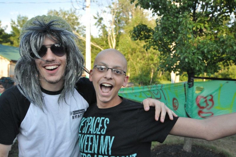 Two people pose while smiling, one wearing a wig.